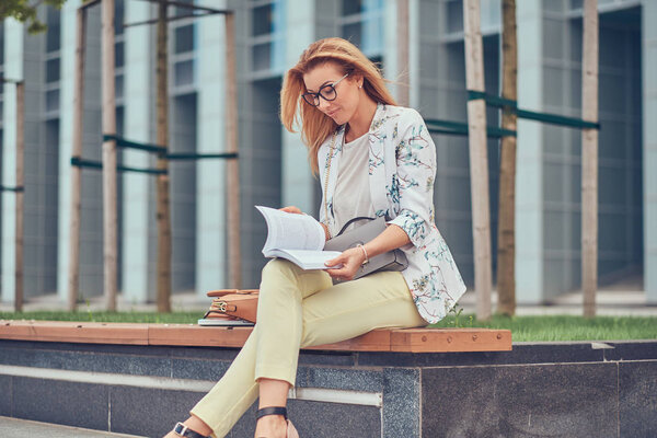 Очаровательная блондинка в современной одежде, учится с книгой, сидит на скамейке в парке против небоскреба
.