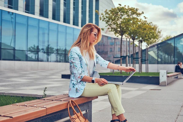 Elegante blogger de moda usando el portátil para trabajar mientras está sentado afuera en un banco contra un rascacielos . —  Fotos de Stock