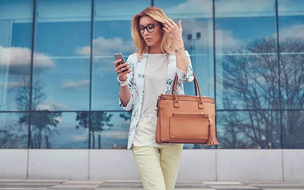 Modische Bloggerin in stylischer Kleidung und Brille mit Handtasche, hält ein Smartphone vor einem Wolkenkratzer. — Stockfoto