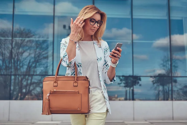 La blogger femminile alla moda in vestiti eleganti e occhiali con una borsa, tiene uno smartphone, in piedi contro un grattacielo . — Foto Stock