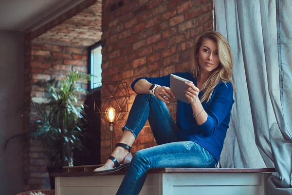 La bloguera rubia de moda sostiene una tableta mientras está sentada en una mesa contra una pared de ladrillo en un estudio con un interior de loft . —  Fotos de Stock