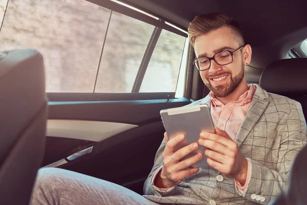 Successful stylish young businessman in a gray suit and pink shirt, riding on a back seat in a luxury car. — Stock Photo, Image