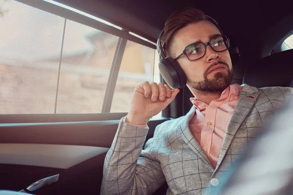 Successful stylish young businessman in a gray suit and pink shirt, riding on a back seat in a luxury car. — Stock Photo, Image