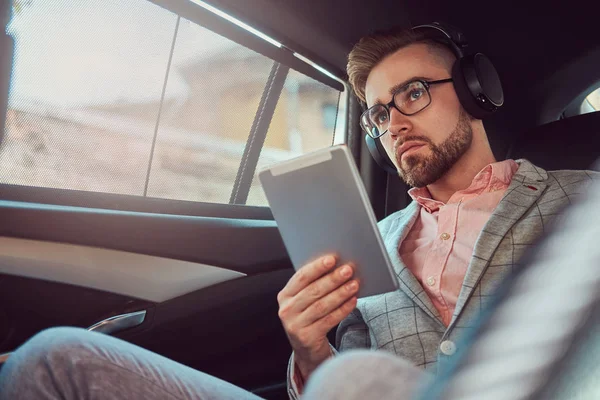 Successful stylish young businessman in a gray suit and pink shirt, riding on a back seat in a luxury car. — Stock Photo, Image