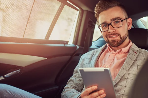 Successful stylish young businessman in a gray suit and pink shirt, riding on a back seat in a luxury car. — Stock Photo, Image