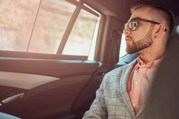 Joven empresario con estilo de éxito en un traje gris y camisa rosa, montado en un asiento trasero en un coche de lujo . — Foto de Stock