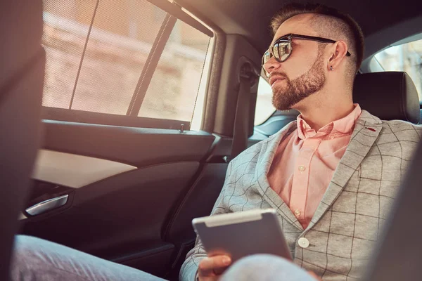 Jovem empresário bem sucedido e elegante em um terno cinza e camisa rosa, montando em um banco de trás em um carro de luxo . — Fotografia de Stock