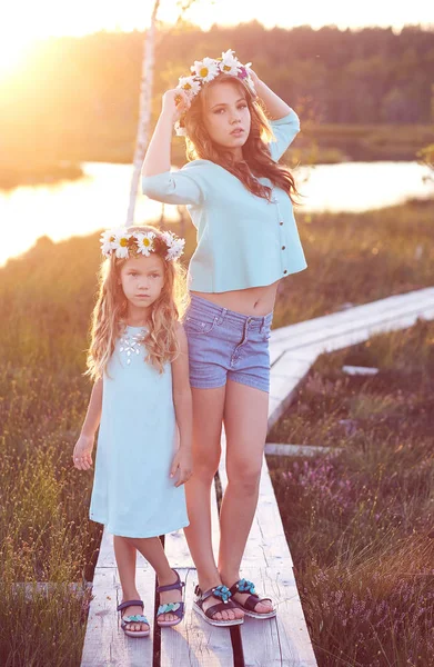 Two beautiful sisters standing against the background of a beautiful landscape, walk on the field near a pond at sunset. — Stock Photo, Image