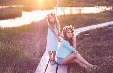 Güzel bir manzara arka plan karşı duran iki güzel kız yürümek bir gölet yakınındaki sahada günbatımında.