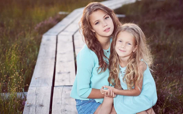 Sisters relaxing outdoors while sitting on a wooden path in a field at sunset. — Stock Photo, Image