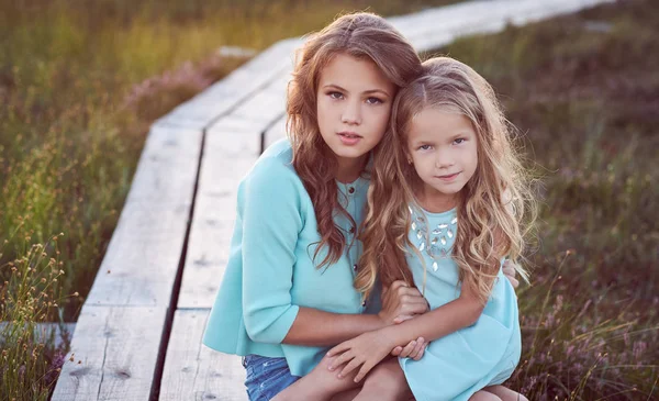 Sisters relaxing outdoors while sitting on a wooden path in a field at sunset. — Stock Photo, Image