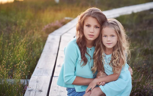 Sisters relaxing outdoors while sitting on a wooden path in a field at sunset. — Stock Photo, Image