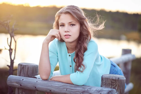 Retrato de una hermosa chica, de pie al aire libre mientras se apoya en una cerca de madera . — Foto de Stock