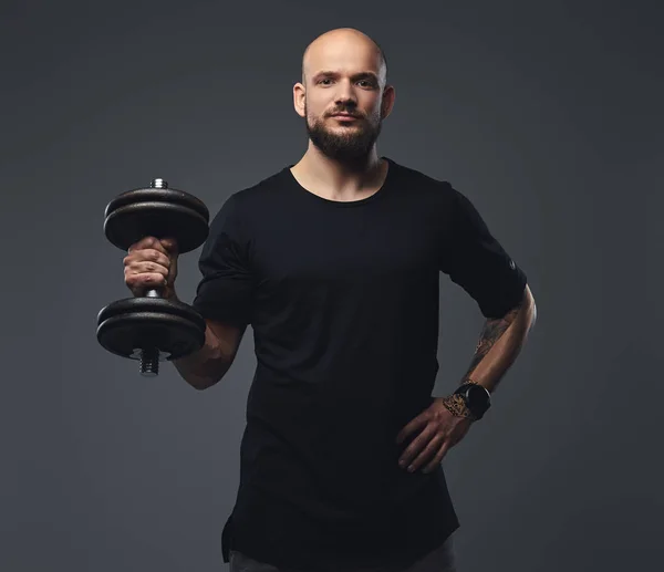 Close-up portrait of a handsome bearded athlete in a black t-shirt, posing dumbbell in a studio.