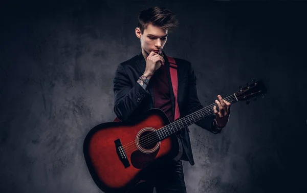 Knappe jonge doordachte musicus met stijlvolle haren in elegante kleding poseren met een gitaar in zijn handen. — Stockfoto