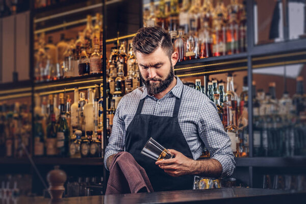 Stylish brutal barman is cleaning the glass with a cloth at bar counter background.