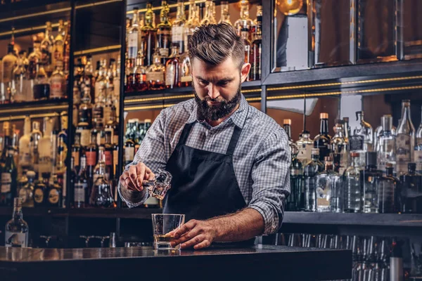 Elegante barman brutal en una camisa y delantal hace un cóctel en el fondo del mostrador del bar . —  Fotos de Stock