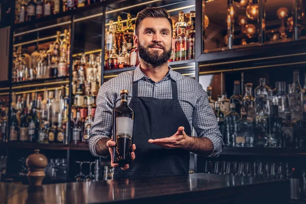 Bir gömlek ve önlük şık acımasız Barmen özel alkol counter arka plan bir şişe sunar. — Stok fotoğraf