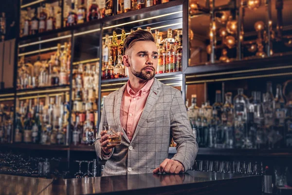 Elegante hombre guapo en un traje elegante sostiene un vaso de whisky en el fondo del mostrador de bar . —  Fotos de Stock