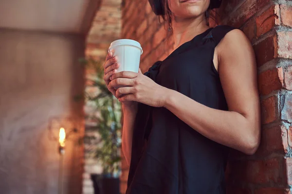 Brunette female in black blouse listening music in headphones and holds a cup of takeaway coffee while leaning against a brick wall in a room with a loft interior.
