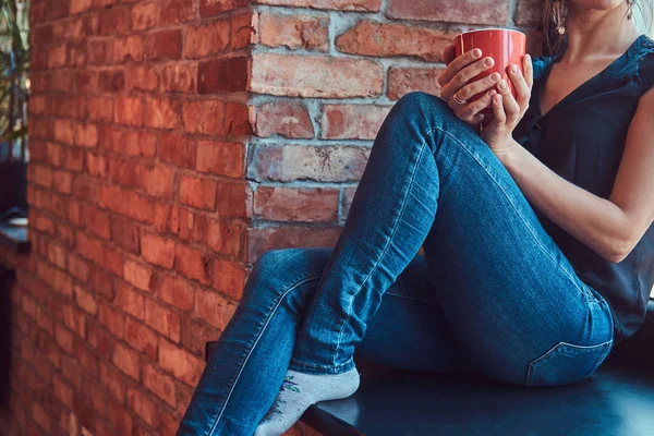 Brunett kvinna i svart blus lyssnar musik i hörlurar och innehar en kopp kaffe medan du sitter på en fönsterbräda i ett rum med loft interiör. — Stockfoto