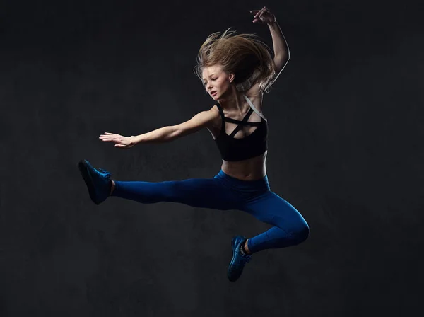 Giovane ballerina bionda in balli di abbigliamento sportivo e salta in uno studio. Isolato su uno sfondo scuro . — Foto Stock