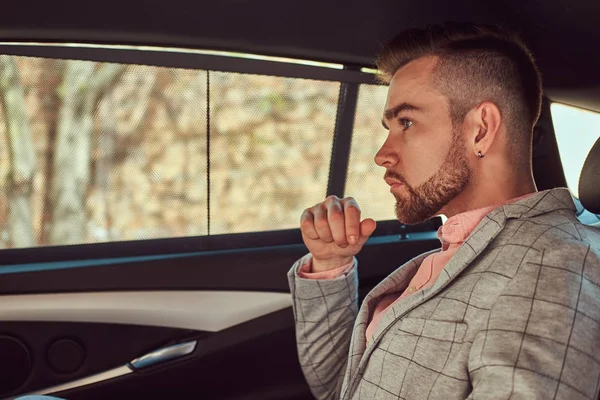 Joven empresario con estilo de éxito en un traje gris y camisa rosa, montado en un asiento trasero en un coche de lujo . — Foto de Stock