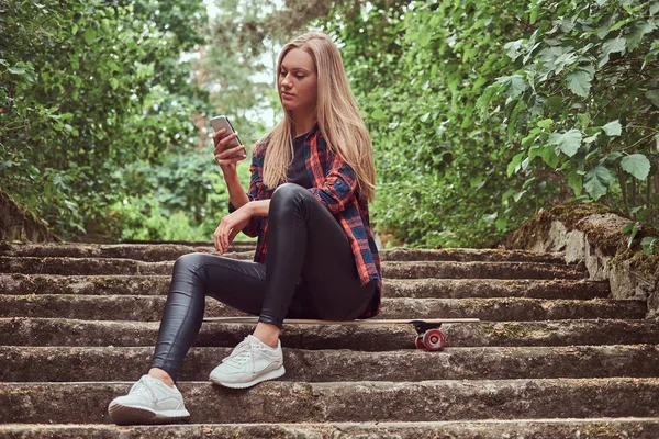 Ung blond hipster flicka med en smartphone medan du sitter på stegen i en park. — Stockfoto