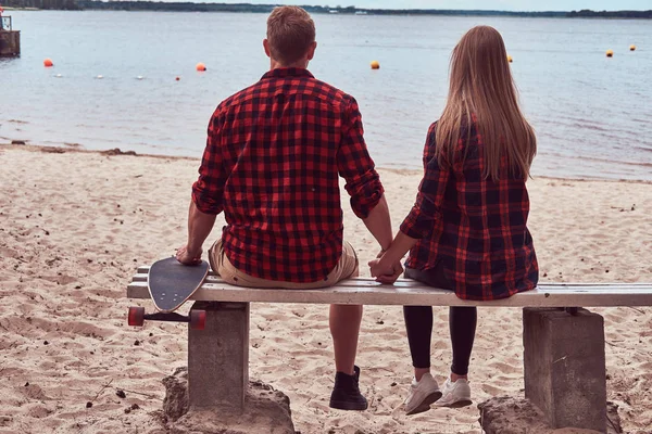 Baksidan på en söt hipster par, sitter på en bänk mitt i stranden och titta på ett hav. — Stockfoto