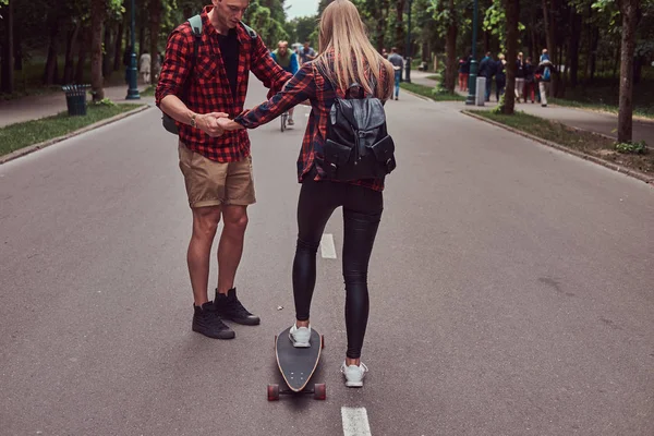年轻的时髦情侣。他在公园的街道上教他的女朋友滑板. — 图库照片