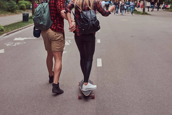Giovane coppia hipster. Guy insegna alla sua ragazza a pattinare per strada in un parco . — Foto Stock