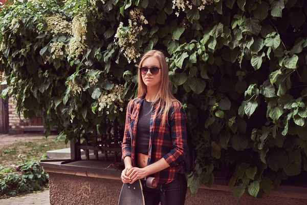 Молода хіпстерська блондинка в повсякденному одязі тримає довгу дошку, стоячи в міському парку . — стокове фото