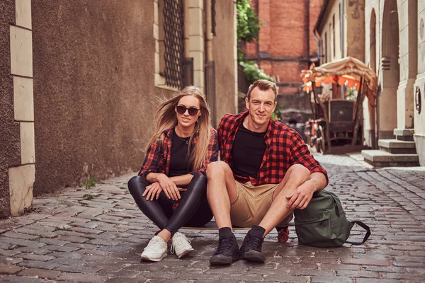 Unga glada hipster par, vila under en promenad som sitter på trottoaren i gamla Europa gatan. — Stockfoto