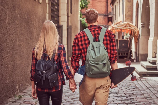 Baksidan på en ung hipster par, stilig skater och hans flickvän, hålla händer, gå runt en gamla smala gatorna i Europa. — Stockfoto