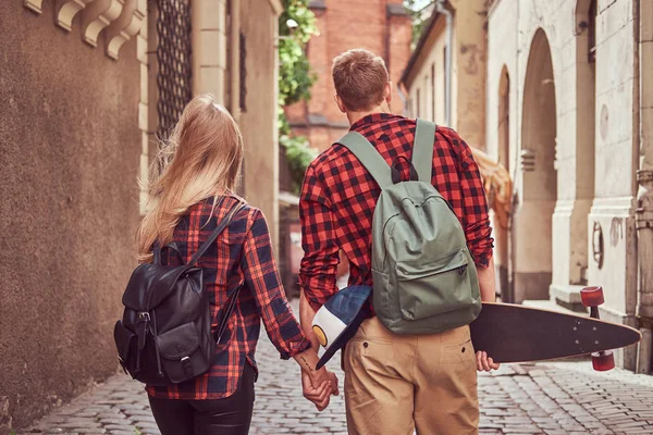 Baksidan på en ung hipster par, stilig skater och hans flickvän, hålla händer, gå runt en gamla smala gatorna i Europa. — Stockfoto