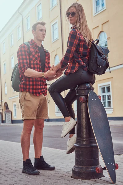 Jonge hipster paar, knappe schaatser hand in hand zijn vriendin die zittend op een brandkraan in een oude Europa straat. — Stockfoto