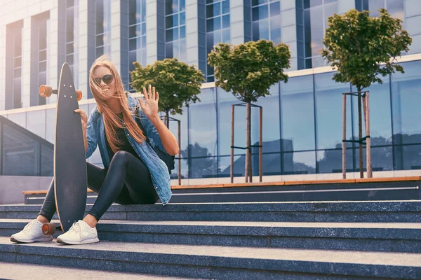 Genç hipster sarışın kız rahat kıyafetler ve adımları bir gökdelen karşı oturup güneş gözlüğü, kaykay üzerinde sürme sonra dinlenme. — Stok fotoğraf