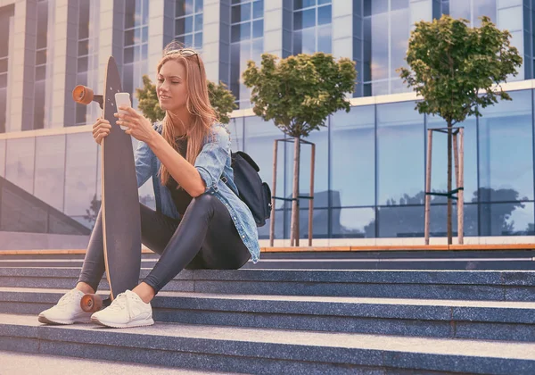 Jonge hipster blond meisje in casual kleding, zittend op stappen tegen een wolkenkrabber, rust na het rijden op het skateboard. — Stockfoto
