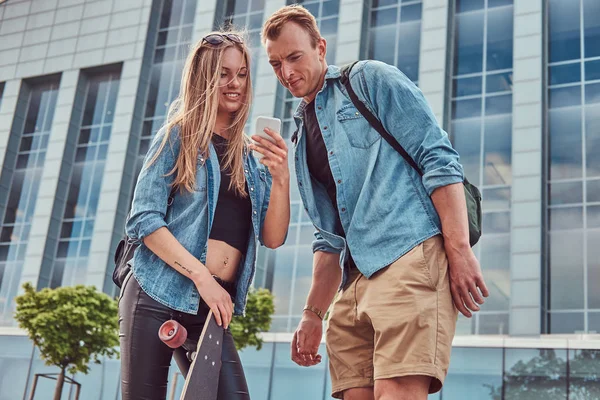 Jonge gelukkige aantrekkelijke paar in casual kleding staan tegen een wolkenkrabber, kijken naar iets op een smartphone. — Stockfoto