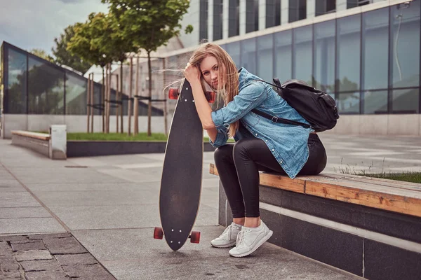 Mladí bokovky blondýnku v neformální clothessitting na lavičce proti mrakodrapu, odpočívá po ježdění na skateboardu. — Stock fotografie