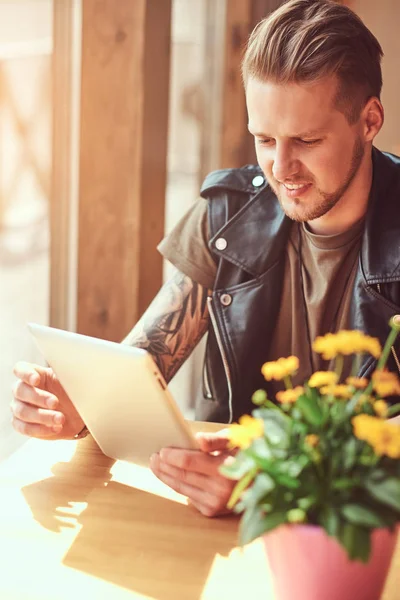 Bello hipster con un taglio di capelli elegante e la barba si siede a un tavolo in un caffè lungo la strada, guarda qualcosa nel computer tablet . — Foto Stock