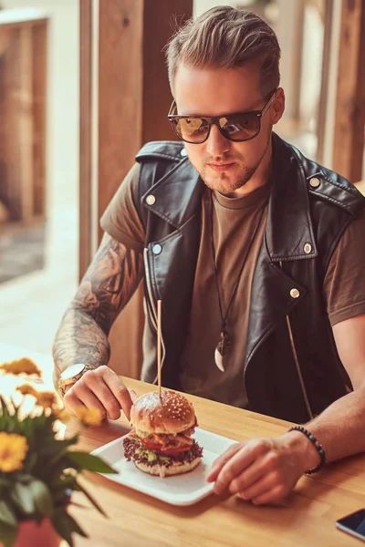 Bonito hipster com um corte de cabelo elegante e barba senta-se a uma mesa em um café à beira da estrada, bebe um refrigerante em um dia quente. . — Fotografia de Stock
