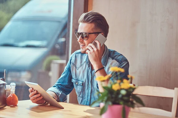 Hipster τύπος με ένα μοντέρνο κούρεμα και γενειάδα κάθεται σε ένα τραπέζι σε ένα καφέ άκρη του δρόμου, μιλώντας στο τηλέφωνο και κατέχει έναν υπολογιστή tablet. — Φωτογραφία Αρχείου