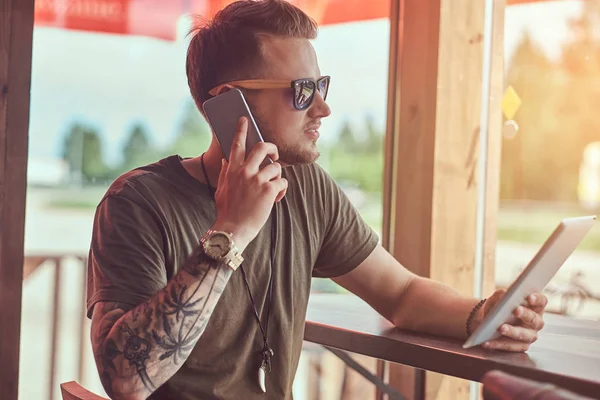 Bonito hipster elegante senta-se a uma mesa em um café à beira da estrada, falando ao telefone e segura o tablet . — Fotografia de Stock