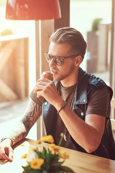 Belle hipster avec une coupe de cheveux élégante et la barbe est assis à une table dans un café en bord de route, boit un soda par une journée chaude . — Photo