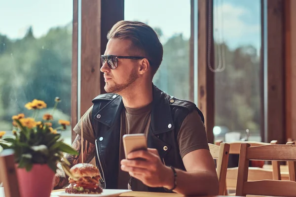 Όμορφος hipster με μοντέρνο κούρεμα και γενειάδα που κάθεται σε ένα τραπέζι, κρατώντας ένα τηλέφωνο κατά τη διάρκεια γεύματος σε οδική καφέ. — Φωτογραφία Αρχείου