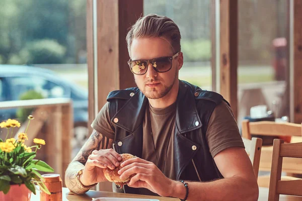 Guapo hipster con un corte de pelo elegante y barba se sienta en una mesa, decidió cenar en un café al lado de la carretera, comiendo una hamburguesa . — Foto de Stock