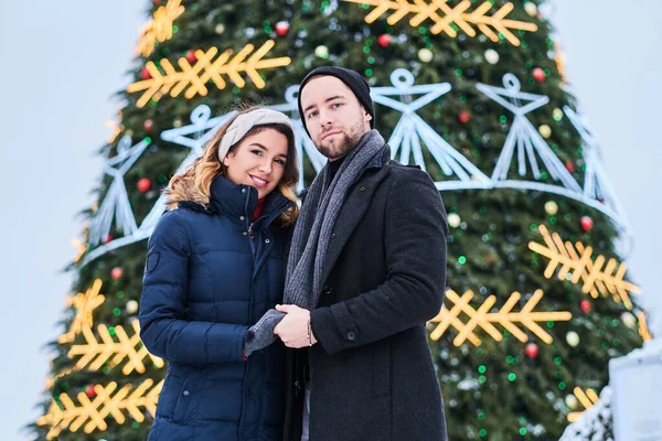 Coppia felice indossando vestiti caldi si tengono per mano e si guardano, in piedi vicino a un albero di Natale della città, godendo di trascorrere del tempo insieme . — Foto Stock