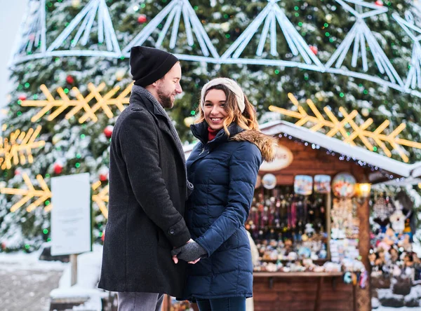 Casal jovem vestindo roupas quentes de mãos dadas e olhar uns para os outros, de pé perto de uma árvore de Natal da cidade, gostando de passar o tempo juntos . — Fotografia de Stock