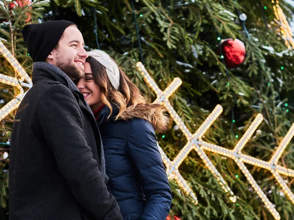 Pareja joven con ropa de abrigo cogida de la mano y mirándose, de pie cerca de un árbol de Navidad de la ciudad, disfrutando pasar tiempo juntos . — Foto de Stock